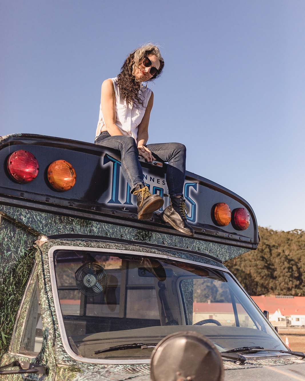 Author Heather Jacks sitting confidently on the roof of a customized black and green bus named Bubba. She wears sunglasses and black boots, perched above the windshield against a clear blue sky, capturing the spirit of nomadic life and independence.