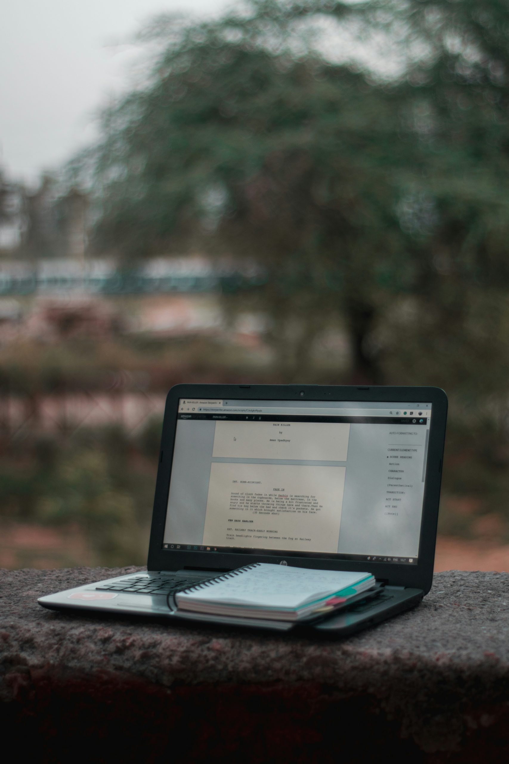 A close-up shot of an open laptop resting on a stone ledge. The screen shows a screenplay formatted in Final Draft software. A small spiral notepad is stacked on the keyboard, with a blurred natural background of trees and a park.