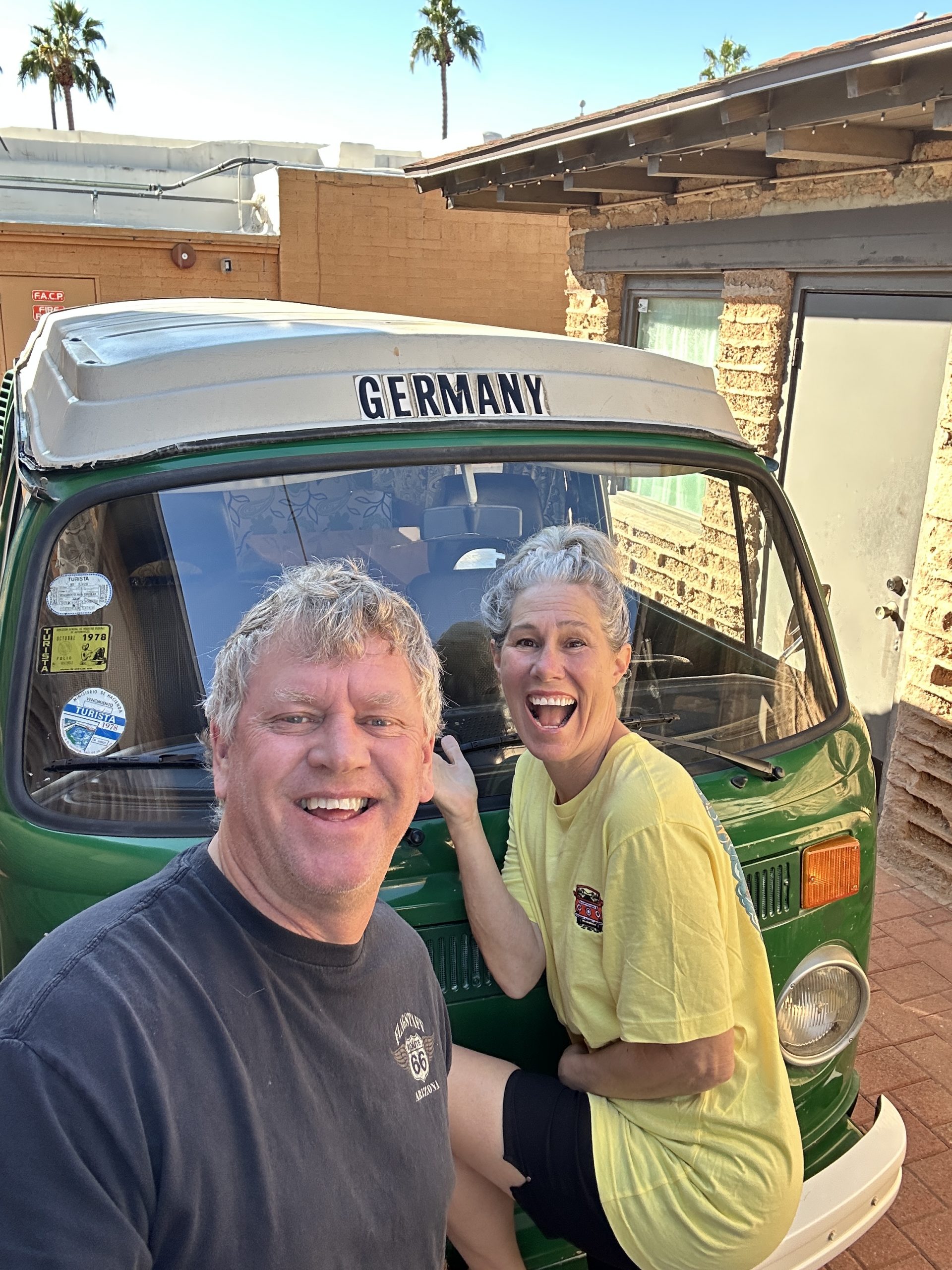 A joyful selfie of Heather and Chris leaning against Buster, their Sumatra Green 1973 VW Bus A smiling man and woman pose excitedly in front of a vintage green Volkswagen bus with a "GERMANY" sign on the pop-top, set against a sunny backdrop with palm trees.