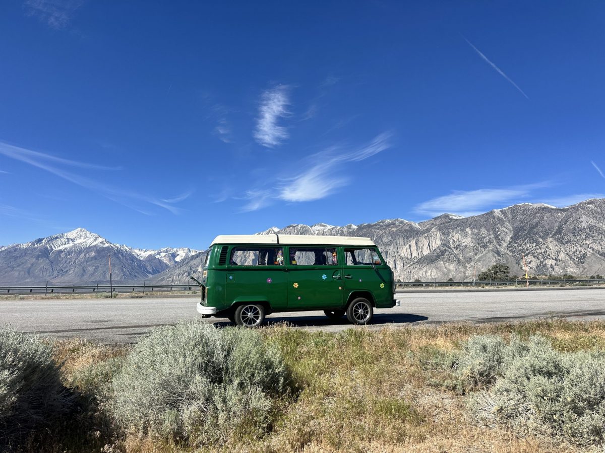 A vintage green and white VW Westfalia camper bus parked on the side of a road, framed by desert sagebrush in the foreground and a massive backdrop of snow-capped mountains under a bright blue sky.