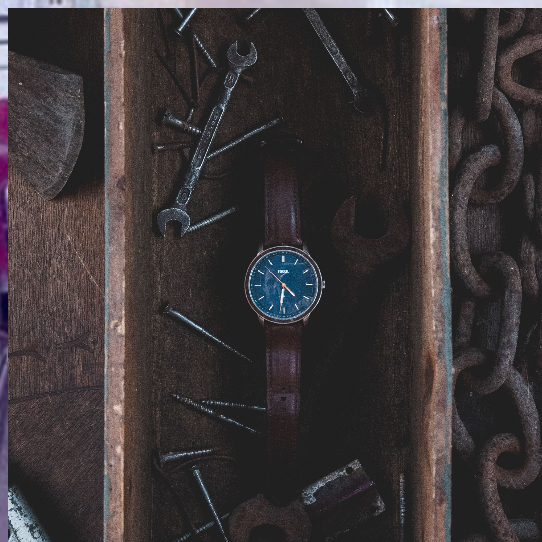 Top-down view of a classic analogue wristwatch with a blue dial, stainless steel casing, and a worn brown leather strap, resting inside a dark wooden box alongside metal tools and chains.